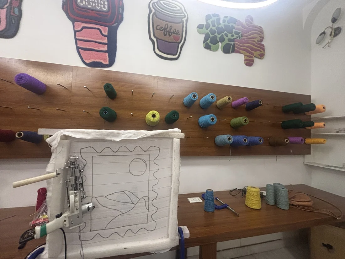 A hand held tufting tool working on a stretched cloth frame, with spools of colored yarn on a wall behind