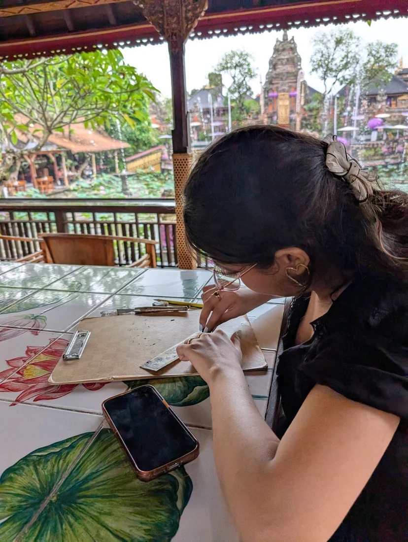 A person working on a craft piece at a table with a view of a Bali style temple garden